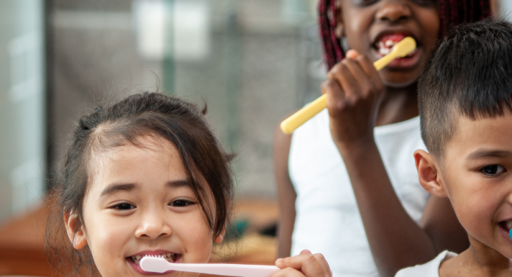 three children brush their teeth looking at the camera