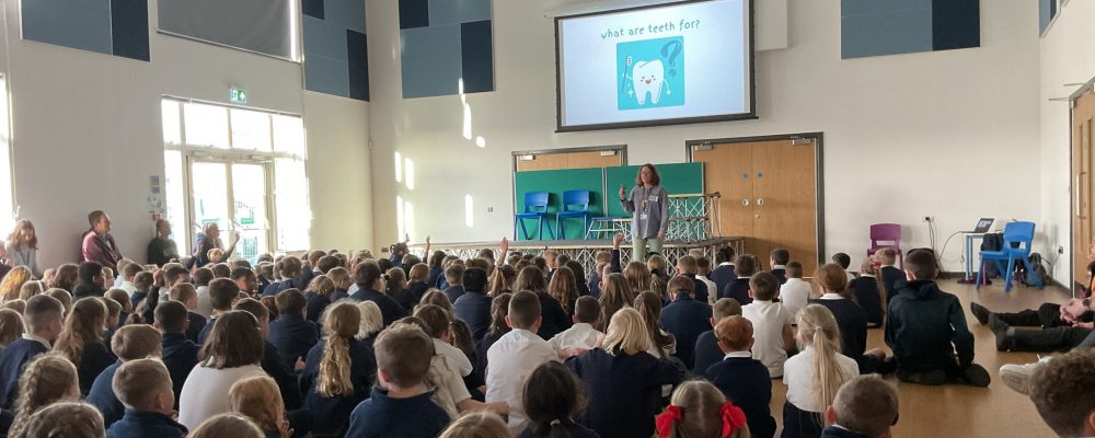 A school hall filled with children with a presenter at the front. On the screen reads "What are teeth for?" with a picture of a tooth