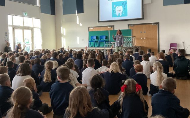 A school hall filled with children with a presenter at the front. On the screen reads "What are teeth for?" with a picture of a tooth